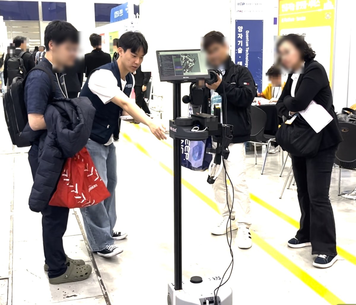 로봇 화면을 손으로 짚어 가며 관람객에게 설명하는 장면. 옆에서 다른 관람객이 카메라로 촬영하고 있다